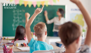 Kids raising hands in classroom with teacher in background