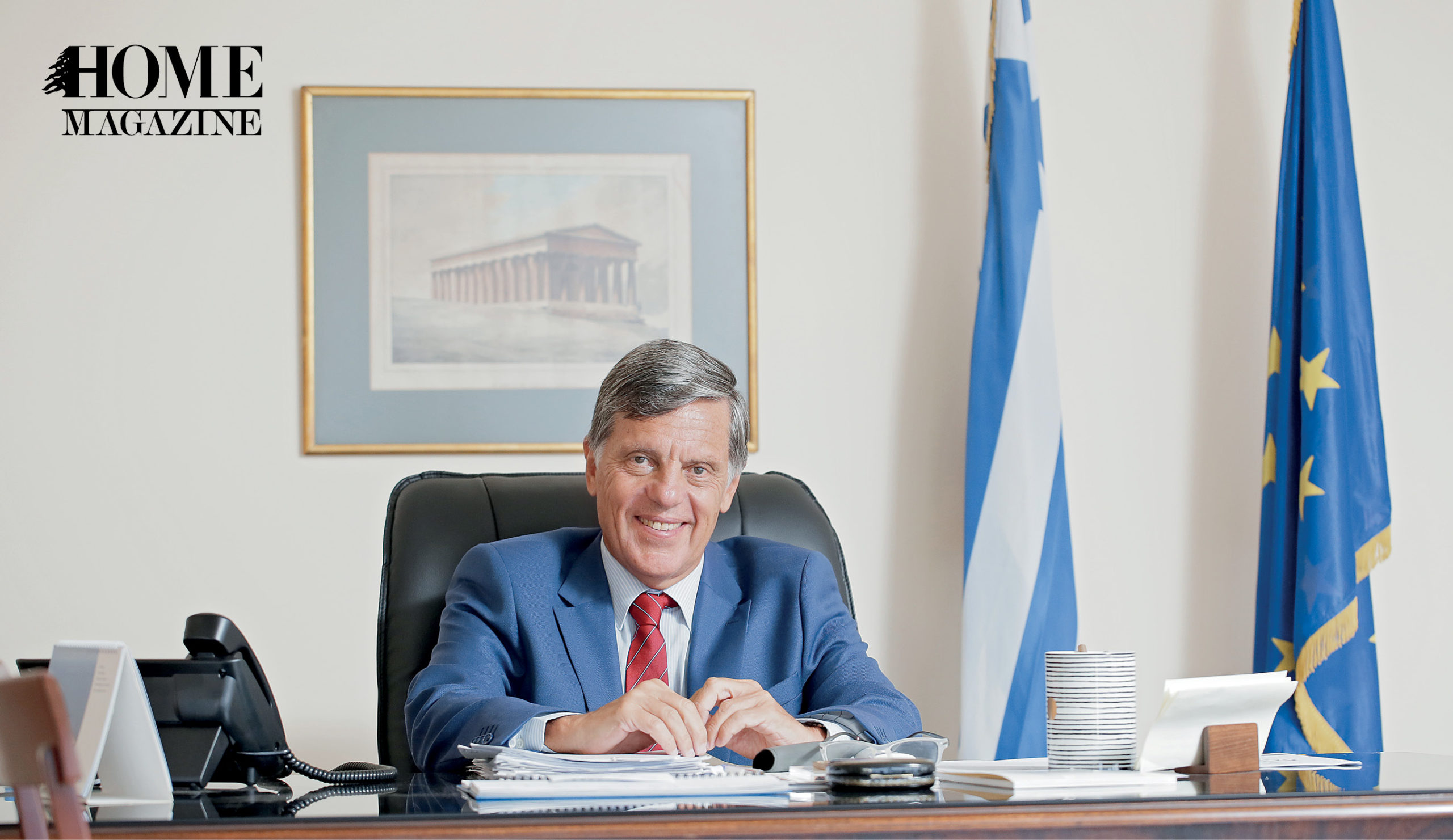 Smiling man wearing navy suit and red tie sitting on desk with background of a picture on wall and two blue flags
