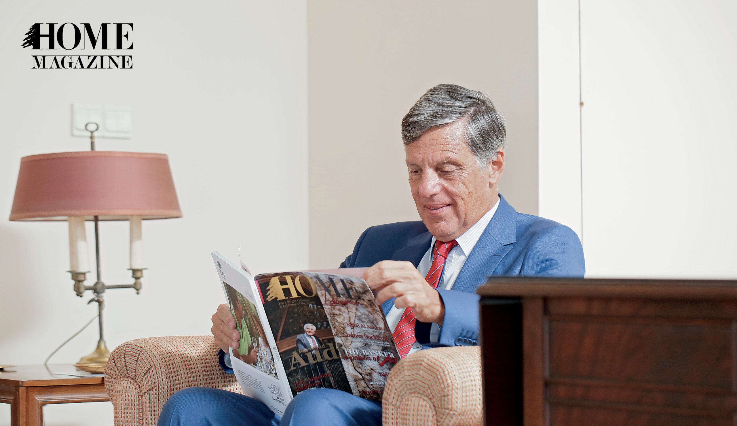 Man wearing navy suit and red tie reading a magazine on chair