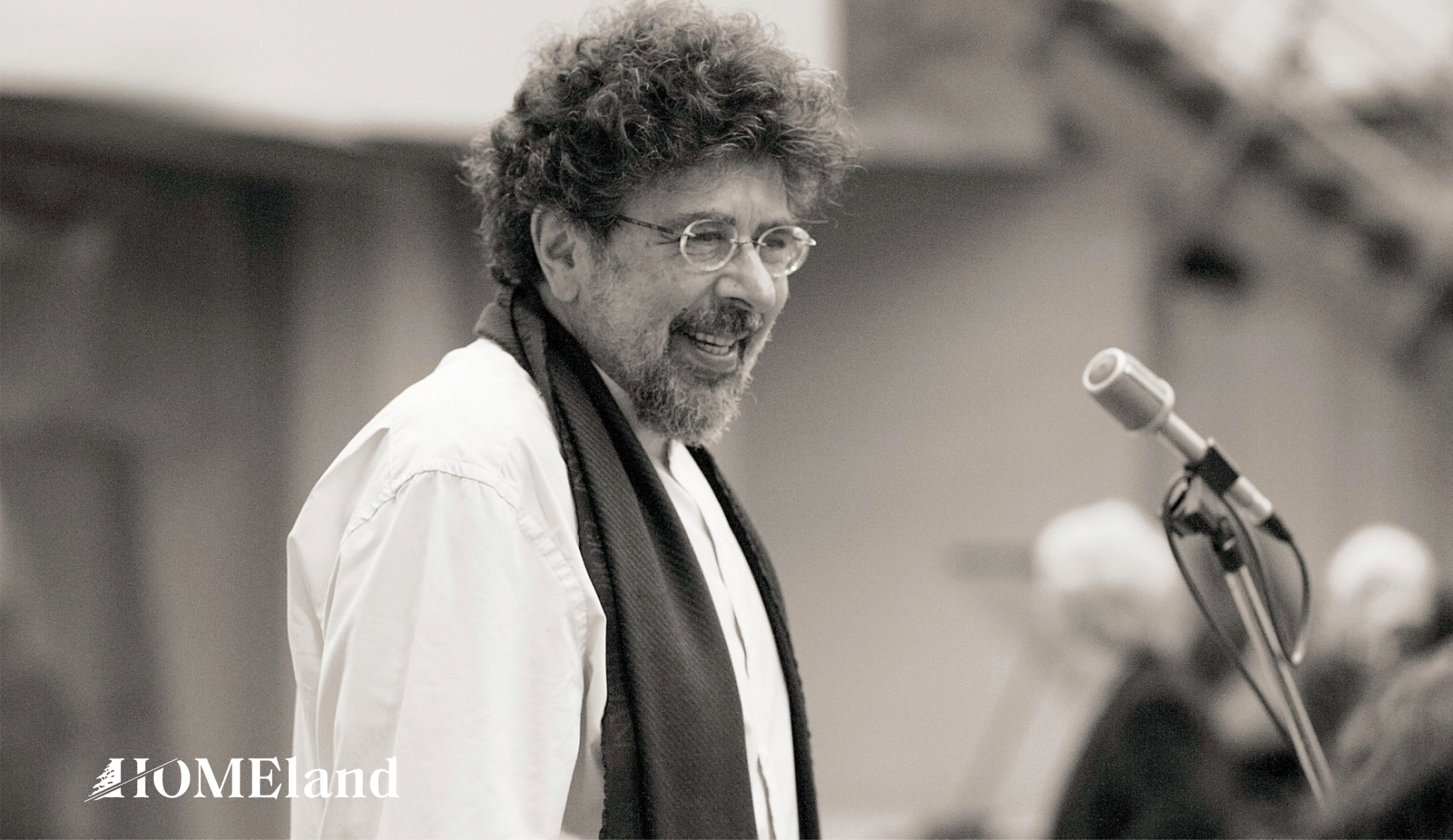 Man wearing white shirt, scarf and eyeglasses standing in front of microphone