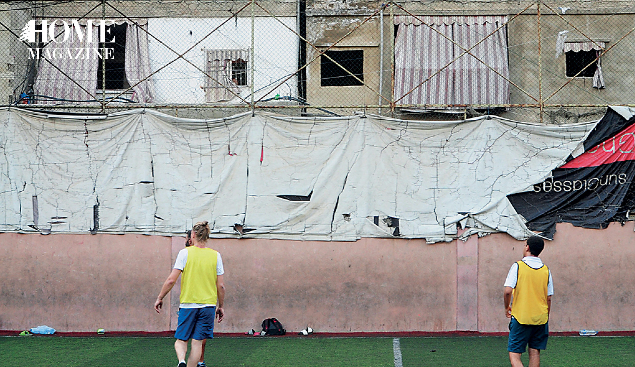 2 boys in blue short and yellow shirt on football court with building behind
