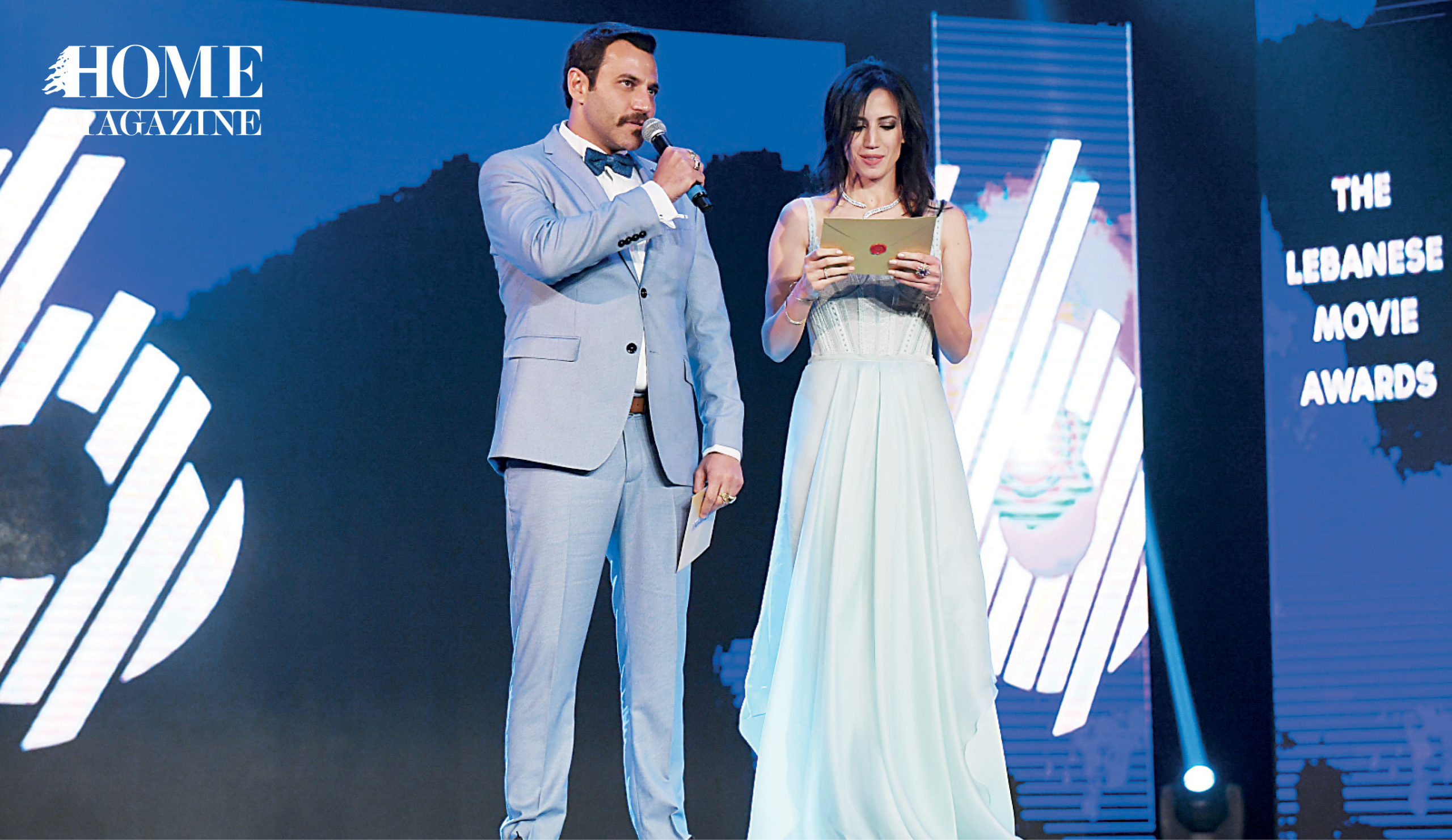 On stage, a woman in light blue dress holding cards and a man in grey suit giving speech on microphone