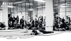 Black and white picture of woman lying on mat floor with microphone in room and crowd behind her