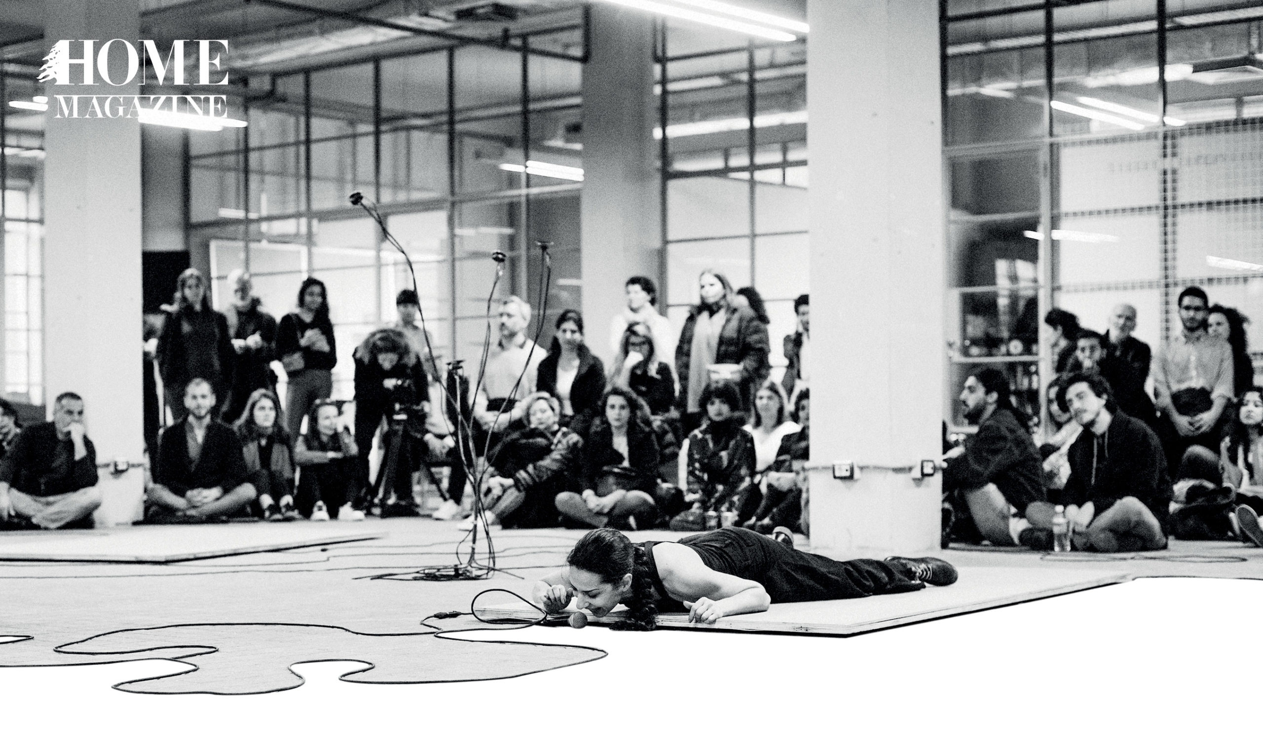 Black and white picture of woman lying on mat floor with microphone in room and crowd behind her